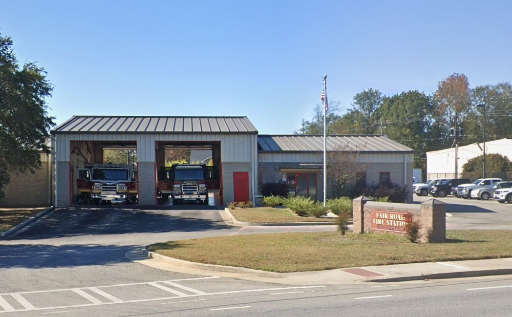 Gray Building with Red Trim and Two Fire Engines Parked in the Bay Garage with a flag pole and sign out front that says "Fair Road Fire Station"